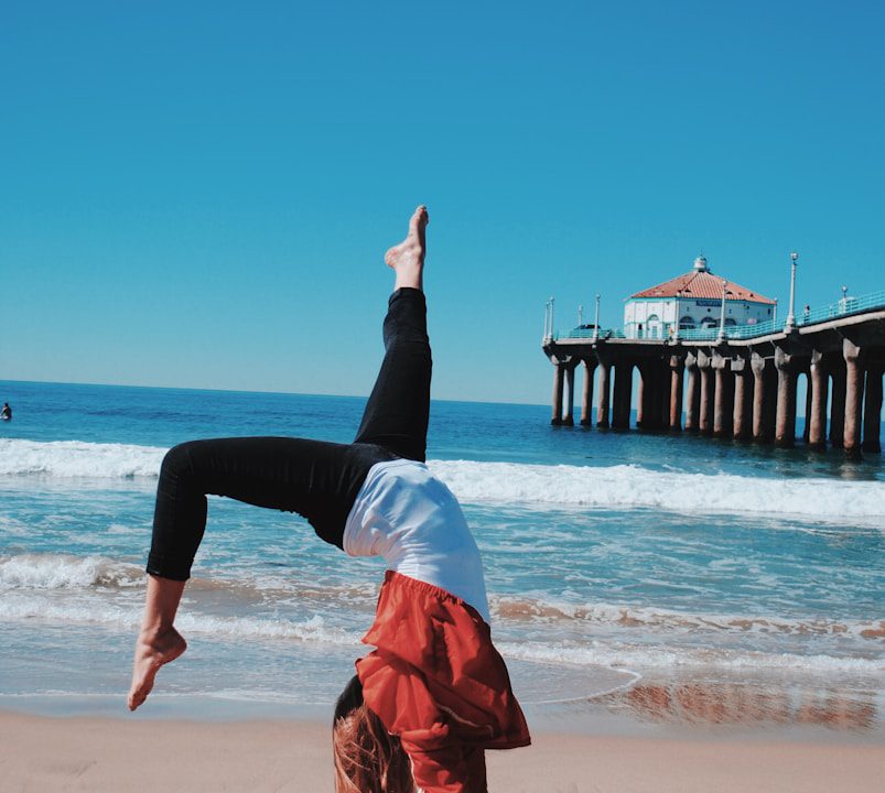 woman wearing red jacket and black leggings doing hand stand in seashore